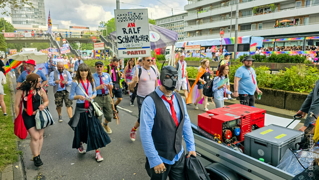 Teil unserer Fußgruppe auf dem CSD 2024 in Köln. Im Vordergrund trägt jemand einen PARTEI-Anzug und eine Puppy-Maske. Im Hintergrund hält jemand ein Schild hoch, auf dem steht: 